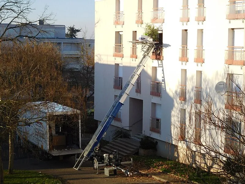 Grue élévatrice devant un immeuble, prête à accéder à un étage pour travaux ou déménagement, au soleil. Déménagement démarches intégrées.