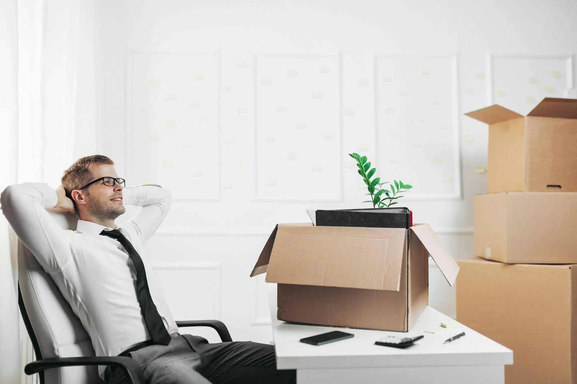 Homme d’affaires détendu dans un fauteuil, entouré de cartons et d’un vase avec une plante sur une table, ambiance déménagement.